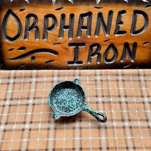 May include: A small, cast iron pan with a turquoise speckled finish. The pan has a handle and two small side handles. The pan is on a brown and white plaid patterned surface. A wooden sign with the words "ORPHANED IRON" is in the background.