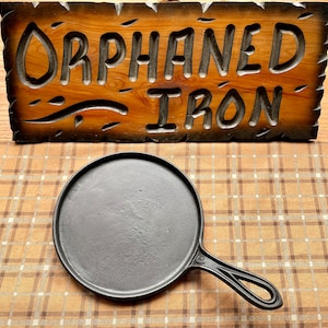May include: A black cast iron skillet with a round cooking surface and a long handle. The skillet is sitting on a brown and white checkered tablecloth. A wooden sign with the words "Orphaned Iron" is in the background.