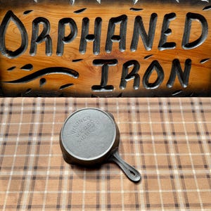 May include: A small, cast iron skillet with a handle, resting on a brown and white plaid surface. The skillet is marked with the text "Wagner Ware Sidney". A wooden sign with the words "Orphaned Iron" is in the background.