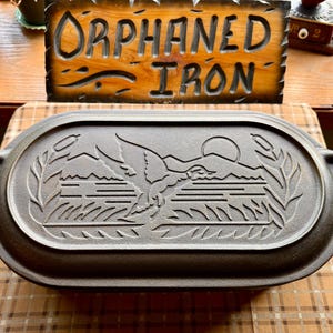 May include: An oval cast iron lid featuring a raised relief design of a duck in flight over water with mountains and a sun in the background, framed by cattails. The lid has two integrated side handles and a dark, matte metallic finish.