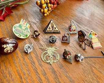May include: A collection of vintage pins and brooches, including a pyramid-shaped pin with "Syrian Shrine" text, a pin with "Kerbella Temple" text, and a pin with a cake design. The pins are arranged on a wooden surface.