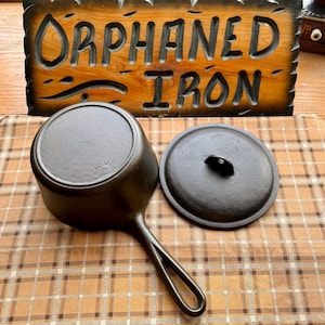 May include: A black cast iron saucepan with a matching lid, featuring a long handle with a teardrop-shaped cutout. The bottom of the pan is embossed with the text 20T. Behind the pan is a wooden sign with the carved text ORPHANED IRON.