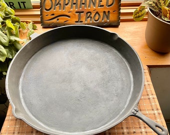 May include: A large, round, cast iron skillet with a black handle. The skillet is sitting on a checkered brown and white surface. The sign above the skillet reads "Orphaned Iron".