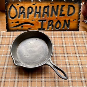 Puede incluir: Una sartén de hierro fundido con un mango largo, sobre una superficie a cuadros marrón y beige. La sartén es de color gris oscuro y parece bien usada. Un letrero de madera con las palabras "ORPHANED IRON" está en el fondo.