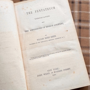 May include: An open book with the title "THE PENTATEUCH VINDICATED" printed in black ink. The book's pages are aged and slightly yellowed, with text detailing the author and publisher. The book is set against a brown and tan plaid background.