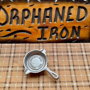 May include: A cast iron ashtray with a handle, featuring the text "COUNTY FAIR SHELBY 1975" in the center. The ashtray is silver and sits on a brown and tan plaid surface. Above it is a wooden sign that reads "ORPHANED IRON".