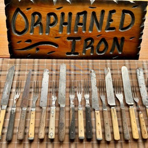 May include: A collection of 18 antique forks and knives with wooden handles. The cutlery is arranged in two rows on a plaid tablecloth. A wooden sign with the words "Orphaned Iron" is visible above the cutlery.