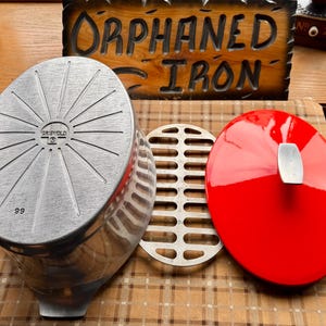 May include: An oval Griswold 99 aluminum roaster with a sunburst pattern on the base, a matching metal rack, and a bright red enameled lid featuring a silver-toned handle. The roaster is positioned next to a wooden sign reading ORPHANED IRON.