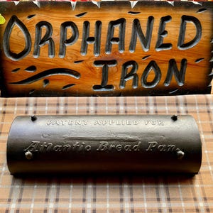 May include: A black cast iron bread pan with the text "Patent Applied For Atlantic Bread Pan" embossed on the front. The pan is sitting on a brown and white checkered surface.