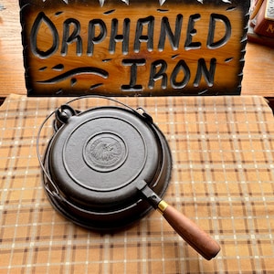 May include: A cast iron waffle iron with a wooden handle. The iron is dark gray and features a circular design with an eagle emblem. The handle is brown and gold. A wooden sign that says "Orphaned Iron" is in the background.