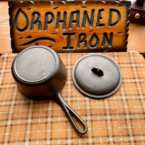 May include: A black cast iron saucepan with a matching lid, featuring a long handle and the text 2QT embossed on the bottom. Behind the pan is a wooden sign with carved text that reads ORPHANED IRON, set against a brown plaid fabric background.
