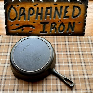 May include: A black cast iron skillet with the text "IRON AGE" and "No. 7" on the bottom. The skillet is sitting on a brown and white plaid surface.