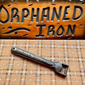 May include: A vintage cast iron stove door opener with the words "Marion Stove Co." embossed on the handle. The opener is lying on a brown and white plaid surface.