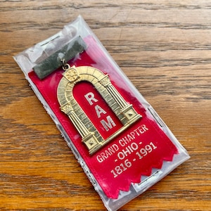 May include: A red ribbon with a gold metal emblem of an archway with the letters "RAM" inside. The ribbon has the text "GRAND CHAPTER - OHIO - 1816 - 1991".