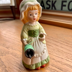 May include: A porcelain figurine of a girl wearing a white bonnet and green dress, holding a spool of green thread. The figurine is a vintage style and has a charming, whimsical look.