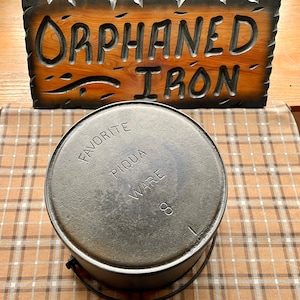 May include: A black cast iron pot with the words "Favorite Piqua Ware" embossed on the side. The pot is sitting on a plaid tablecloth.