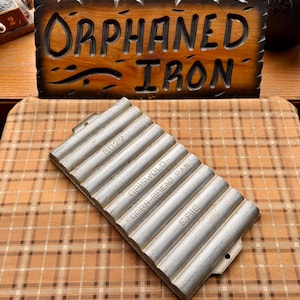 May include: A vintage Griswold cast iron corn bread pan with a ribbed design. The pan is silver and has the text "8022", "Griswold", "Corn Bread Pan", and "Erie" embossed on it. A wooden sign that says "Orphaned Iron" is in the background.