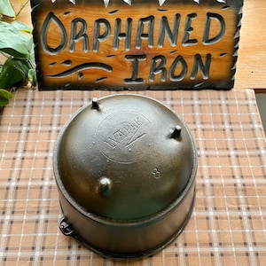 May include: A cast iron Dutch oven with a black finish and a logo that reads "Wapak" with a small arrow. The oven has three legs and a handle. The oven is 8 inches in diameter.