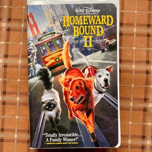 May include: A VHS tape for the Disney movie "Homeward Bound II: Lost in San Francisco." The cover features a colorful illustration of a cat, a golden retriever, and a white dog running on a street with a cable car and the Golden Gate Bridge in the background.