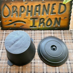 May include: A black cast iron pot with a lid. The pot has a number "4" on the side. The lid has a knob in the center. The pot and lid are sitting on a plaid fabric surface. The words "Orphaned Iron" are carved into a wooden sign in the background.