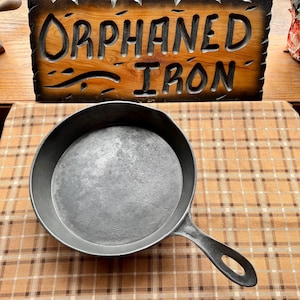 May include: A cast iron skillet with a long handle and a circular cooking surface. The skillet is gray and sits on a brown and tan plaid surface. A wooden sign with the words "ORPHANED IRON" is in the background.