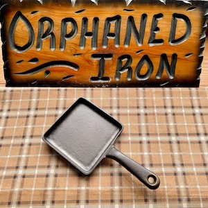 May include: A square cast iron skillet with a single handle, featuring a textured black surface. The pan is positioned in front of a wooden sign with carved, dark-stained lettering that reads ORPHANED IRON, set against a brown plaid fabric.