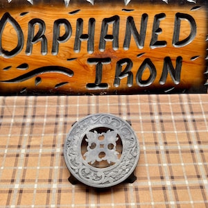 May include: A wooden sign with the words "ORPHANED IRON" carved into it, above a decorative metal trivet. The sign is brown with black lettering and a decorative edge. The trivet is gray and has an ornate design.