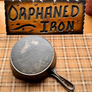 May include: A vintage cast iron skillet with a handle, resting on a plaid surface. The skillet has the text "SIDNEY HOLLOW WARE CO." and the number "5" embossed on the bottom. A wooden sign with the words "ORPHANED IRON" is in the background.
