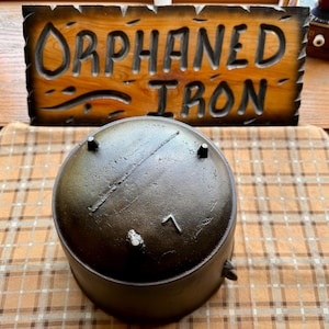 May include: A dark, cast iron pot with a lid, featuring three small feet and a handle. The pot is set against a backdrop of a wooden sign that reads "ORPHANED IRON". The sign is carved into the wood.
