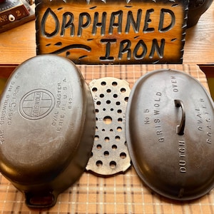 May include: An antique Griswold cast iron oval roaster set, including the Dutch oven lid, the oval base, and a perforated metal insert. The set is brown and features the Griswold logo and text. The sign in the background reads "Orphaned Iron."