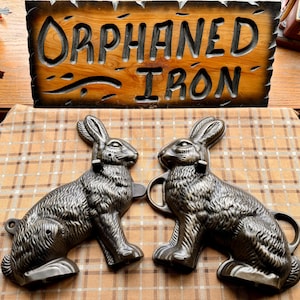 May include: Two cast iron rabbit molds with handles, displayed on a plaid surface. The molds are detailed with fur texture and have a dark metallic finish. A wooden sign with the words "Orphaned Iron" is in the background.