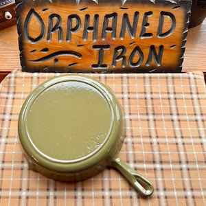 May include: An olive green cast iron skillet with a long handle. The skillet is sitting on a brown and tan plaid tablecloth. A wooden sign that reads "ORPHANED IRON" is in the background.