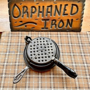 May include: A vintage cast iron waffle iron with a black handle and a waffle pattern on the cooking surface. The waffle iron is on a checkered tablecloth.