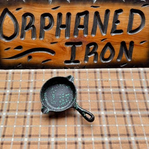 May include: A small, black cast iron skillet with a handle, speckled with green paint. The skillet sits on a brown and tan plaid surface, with a wooden sign that reads "Orphaned Iron" in the background.