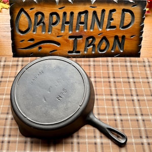 May include: A cast iron skillet with the words "Iron Age" and "No. 8" stamped on the bottom. The skillet is sitting on a brown and white checkered tablecloth.