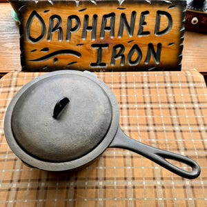 May include: A round black cast iron skillet with a matching lid and a long handle. Behind the skillet is a rectangular wooden sign with carved, dark-stained lettering that reads ORPHANED IRON, featuring a natural wood grain finish.