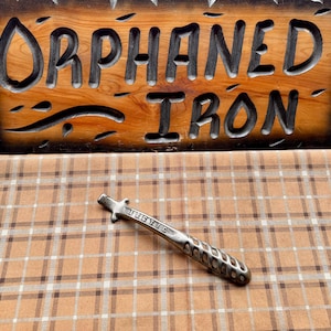 May include: A wooden sign with the words "ORPHANED IRON" carved into it, with a decorative metal object in front. The sign is made of light brown wood with dark brown carved letters. The metal object is silver and has a textured handle.