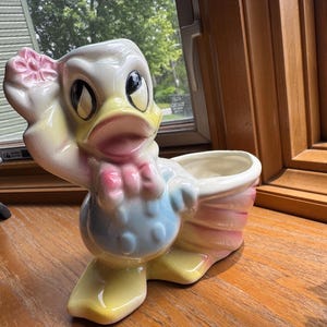 May include: A ceramic duck planter featuring a glossy finish with pastel yellow, blue, and pink accents. The duck has a raised wing holding a pink flower, a wide open beak, and large dark eyes. The body forms a hollow basin for small plants.