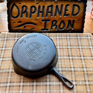 May include: A black cast iron skillet with a handle, featuring the "Griswold" logo and text. The skillet is marked "Cast Iron" and "Skillet" with the city "Erie, PA" visible. The background includes a wooden sign that reads "Orphaned Iron."
