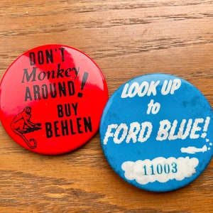 May include: Two round button pins. The red pin has a black monkey with the text "Don't Monkey Around! Buy Behlen". The blue pin has white text that says "Look Up to Ford Blue!" with the number "11003" below.