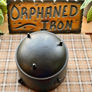 May include: A black cast iron pot with the text "SIDNEY HOLLOWAY SIDNEY 8" on the bottom. The pot is sitting on a checkered surface.