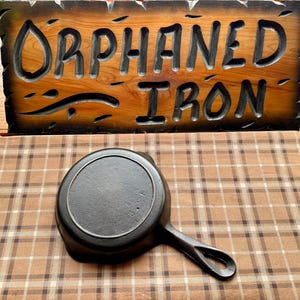 May include: A black cast iron skillet with a handle. The skillet is on a brown and white plaid tablecloth. The text "Orphaned Iron" is visible on a wooden sign in the background.
