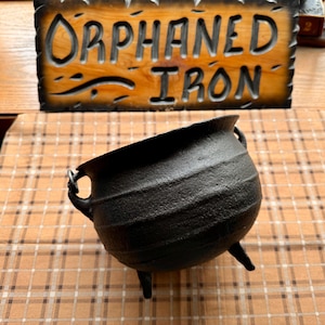 May include: A black cast iron cauldron with a rounded body, three short legs, and two side handles. Behind the cauldron is a rectangular wooden sign with a dark, scalloped edge and the text ORPHANED IRON carved into the light brown wood surface.