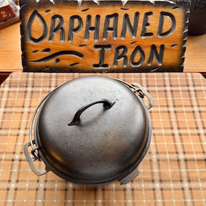 May include: A cast iron Dutch oven with a lid and handle. The oven is dark gray and sits on a checkered surface. A wooden sign with the words "Orphaned Iron" is in the background.