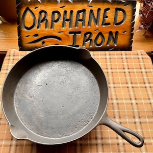 May include: A well-worn, dark gray cast iron skillet with a long handle and a rounded cooking surface. The skillet is sitting on a patterned surface, with a wooden sign that reads "ORPHANED IRON" in the background.