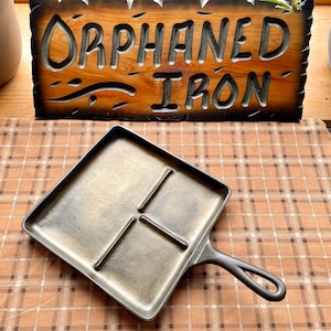 May include: A cast iron skillet with a divided cooking surface. The skillet has a black handle and is sitting on a brown and white checkered tablecloth. A wooden sign with the words "Orphaned Iron" is in the background.