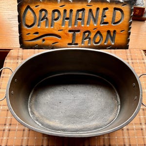 May include: An oval-shaped cast iron roasting pan with two metal loop handles attached by rivets on each side. Behind the pan is a rectangular wooden sign with carved, dark-stained lettering that reads ORPHANED IRON.