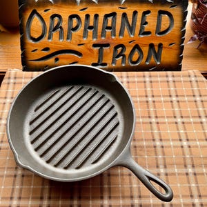 May include: A cast iron grill pan with a handle, featuring a ribbed cooking surface. The pan is dark gray and sits on a checkered brown and tan surface. A wooden sign with the words "Orphaned Iron" is in the background.
