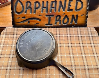 May include: A cast iron skillet with a handle, resting on a plaid surface. The skillet is marked "GRISWOLD'S ERIE" and "699B". A wooden sign with the words "ORPHANED IRON" is in the background.