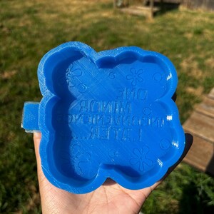 Op de afbeelding: Een helderblauwe, bloemvormige mal met de tekst "ONE MINUTE LATER!" en decoratieve bloemelementen. De mal wordt in een hand gehouden tegen een achtergrond van groen gras en een houten hek.